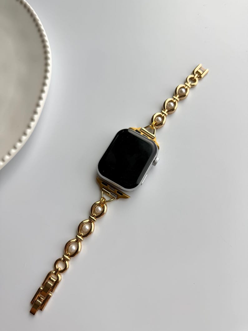 May include: A gold-toned watch band with a pearl accent is attached to a silver smartwatch with a black screen. The band has a chain-like design with oval-shaped pearls. The watch and band are on a white surface.