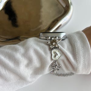 May include: A silver-toned watch band accessory featuring a series of charms including a star with embedded clear crystals, a white heart-shaped charm, and a flower-shaped charm, all connected by a delicate silver-toned metal chain link.