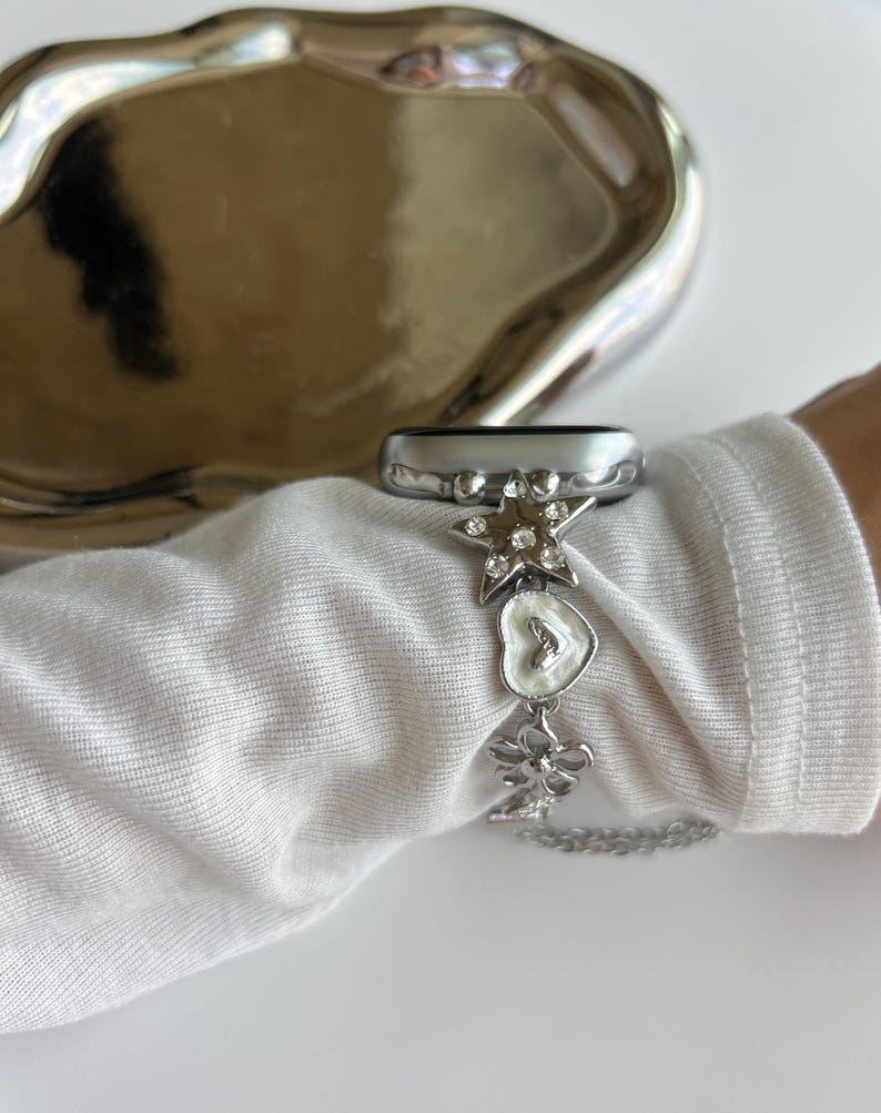 May include: A silver-toned watch band accessory featuring a series of charms including a star with embedded clear crystals, a white heart-shaped charm, and a flower-shaped charm, all connected by a delicate silver-toned metal chain link.