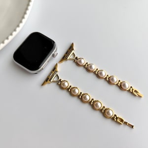 May include: Gold-toned Apple Watch band with faux pearls. The band features a series of round, cream-colored pearls set in a gold metal link design. The watch face is black with a silver frame. The band is unattached.