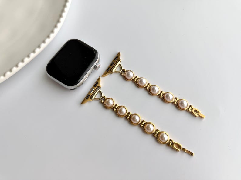 May include: Gold-toned Apple Watch band with faux pearls. The band features a series of round, cream-colored pearls set in a gold metal link design. The watch face is black with a silver frame. The band is unattached.