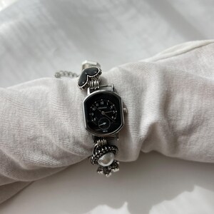 May include: A silver wristwatch with a black, rectangular face. The watch has a decorative chain and embellishments, including a heart-shaped design and a floral charm. The watch face displays the time with white numerals. The watch is a stylish accessory.