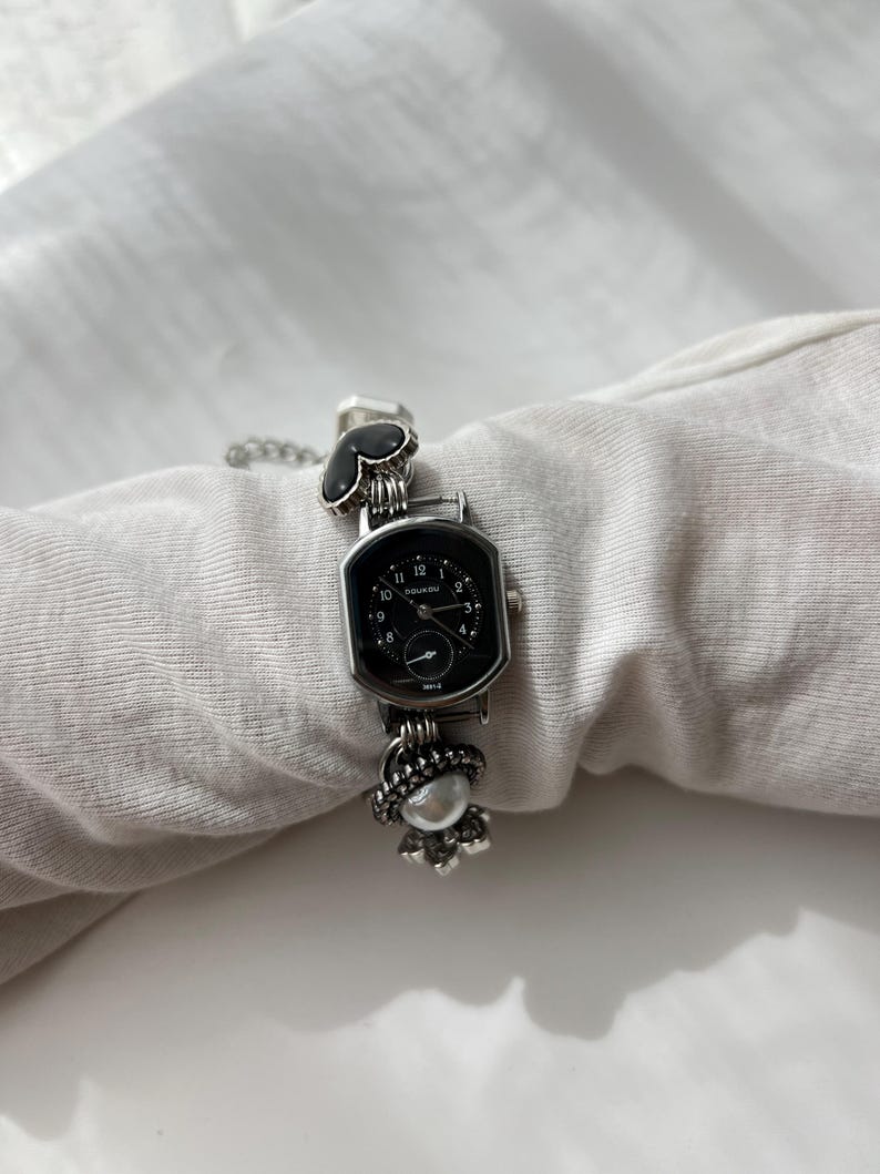May include: A silver wristwatch with a black, rectangular face. The watch has a decorative chain and embellishments, including a heart-shaped design and a floral charm. The watch face displays the time with white numerals. The watch is a stylish accessory.