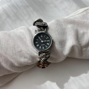 May include: A silver quartz watch with a black face and Roman numeral hour markers. The watch strap has a unique design with decorative links and dark accents. The watch is displayed on a white surface.