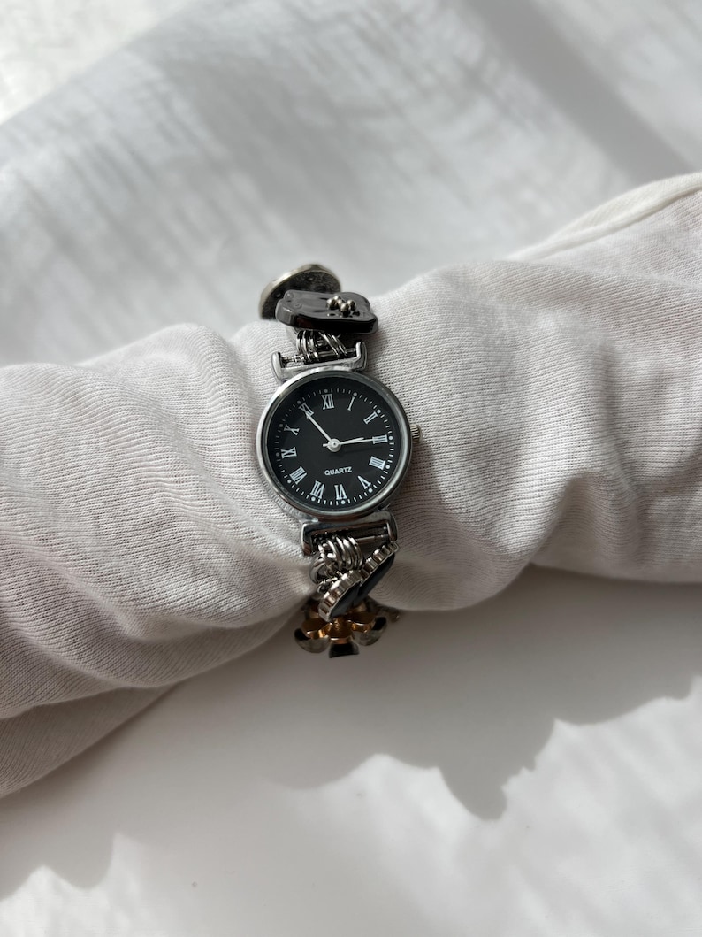 May include: A silver quartz watch with a black face and Roman numeral hour markers. The watch strap has a unique design with decorative links and dark accents. The watch is displayed on a white surface.