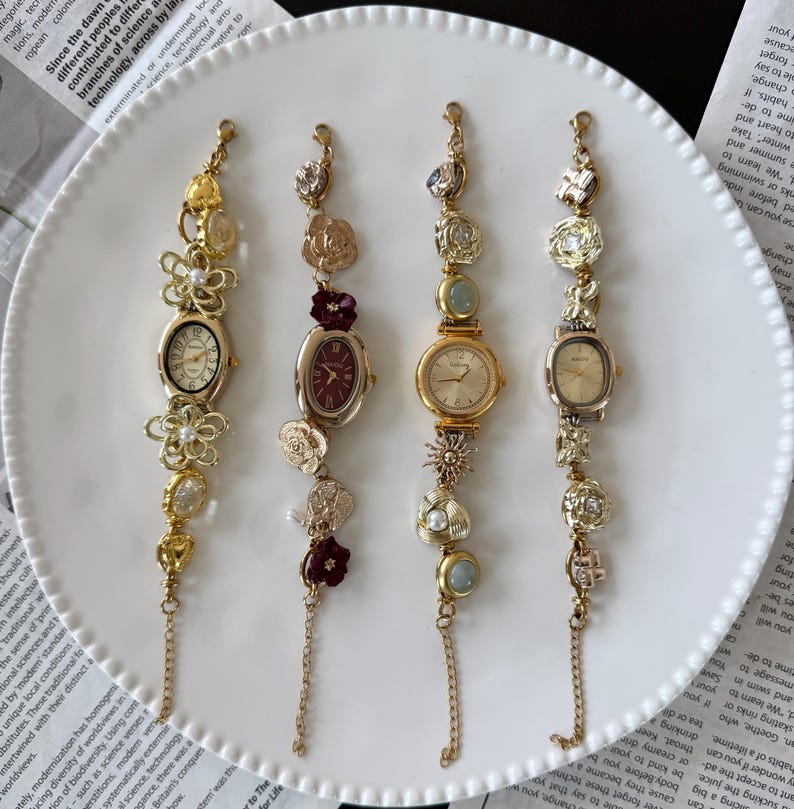 May include: Four vintage-style gold-toned wristwatches with decorative floral and gem accents. Each watch has a different face shape and is adorned with pearls, crystals, and charms. The watches are displayed on a white plate.