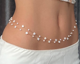 May include: A gold chain body chain with white star-shaped beads and small white pearl beads. The chain is worn around the waist.
