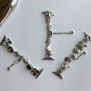 May include: Three silver-toned metal watch bands featuring decorative charms. Each band includes various shapes such as flowers, hearts, stars, and butterflies, accented with small clear crystals and dangling chain elements with heart-shaped ends.
