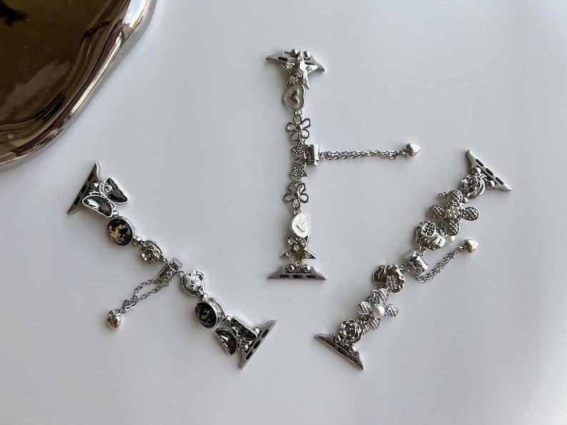 May include: Three silver-toned metal watch bands featuring decorative charms. Each band includes various shapes such as flowers, hearts, stars, and butterflies, accented with small clear crystals and dangling chain elements with heart-shaped ends.