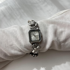 May include: A silver wristwatch with a square face and Roman numeral hour markers. The watch band features ornate floral and geometric designs. The watch face displays the brand name "NAIDU". The watch is displayed on a white surface.