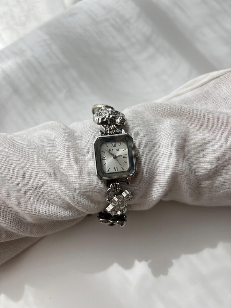 May include: A silver wristwatch with a square face and Roman numeral hour markers. The watch band features ornate floral and geometric designs. The watch face displays the brand name "NAIDU". The watch is displayed on a white surface.