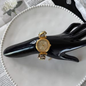 May include: A gold-toned wristwatch with a round face and a light-colored dial. The watch features a decorative band with ornate detailing and is displayed on a black hand-shaped stand. The watch is set against a white plate and newspaper background.