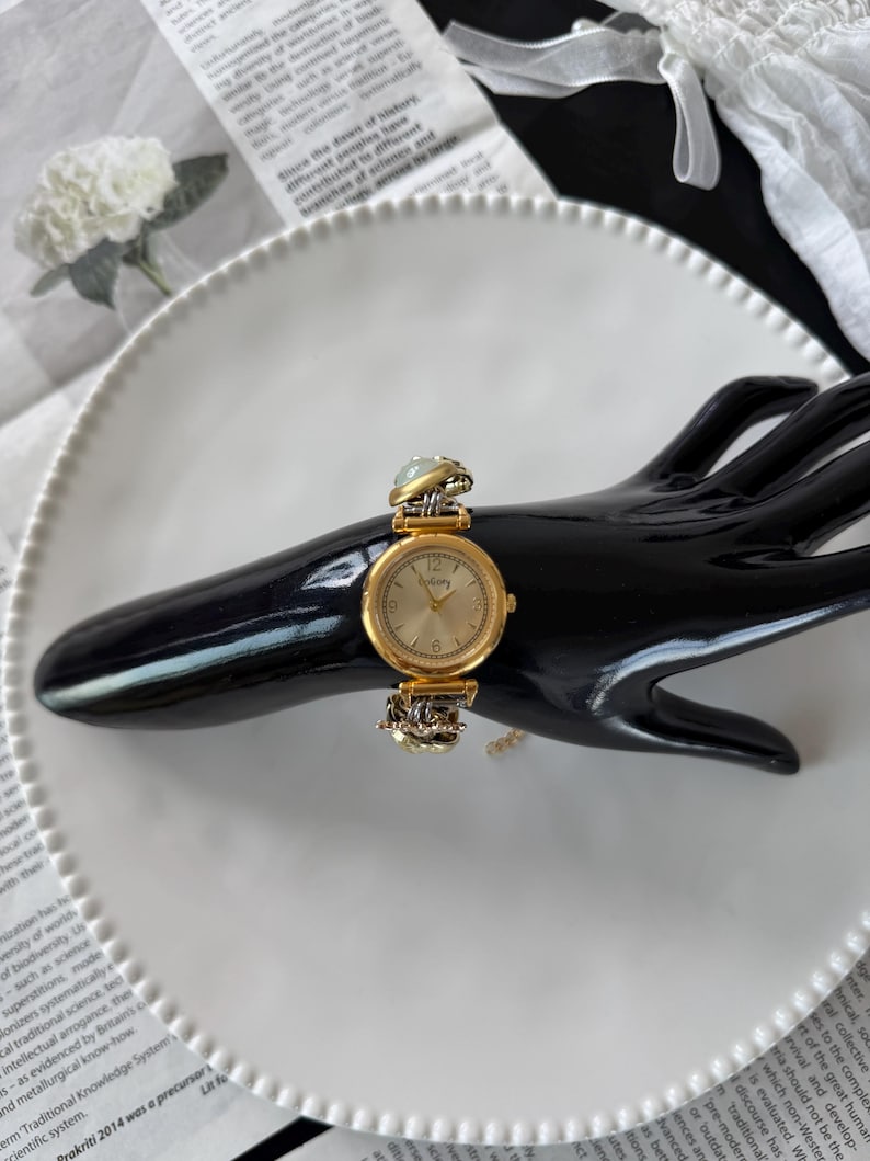 May include: A gold-toned wristwatch with a round face and a light-colored dial. The watch features a decorative band with ornate detailing and is displayed on a black hand-shaped stand. The watch is set against a white plate and newspaper background.
