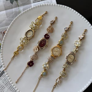 May include: Four decorative vintage-style wristwatches with ornate gold-tone and silver-tone metalwork. Each watch features a different face shape and is adorned with floral and pearl accents. The watches are displayed on a white plate.
