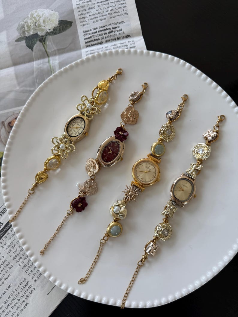 May include: Four decorative vintage-style wristwatches with ornate gold-tone and silver-tone metalwork. Each watch features a different face shape and is adorned with floral and pearl accents. The watches are displayed on a white plate.