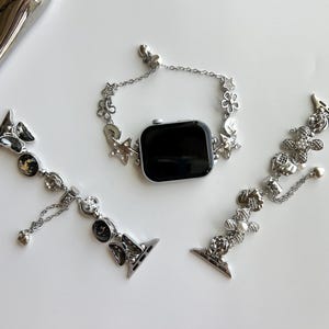 May include: A silver-toned smartwatch with a black screen and two detachable watch bands. The bands feature intricate metal charms including stars, flowers, and circular accents with embedded crystals and pearls, connected by delicate silver chains.