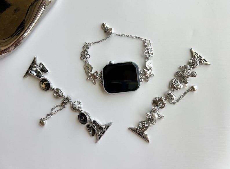 May include: A silver-toned smartwatch with a black screen and two detachable watch bands. The bands feature intricate metal charms including stars, flowers, and circular accents with embedded crystals and pearls, connected by delicate silver chains.