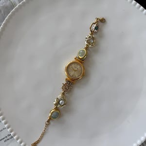 May include: A gold-toned wristwatch with a round face and decorative embellishments. The watch band features various charms, including a pearl, a sunburst, and oval-shaped stones. The watch is displayed on a white plate.