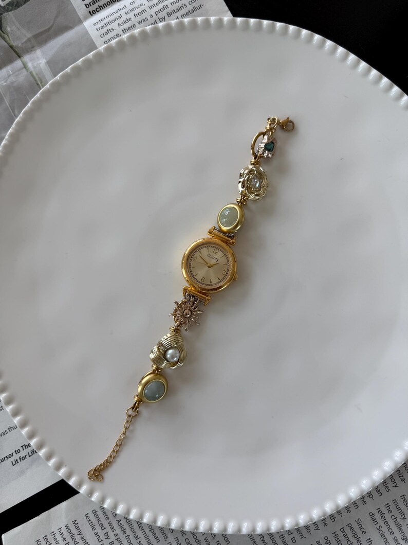 May include: A gold-toned wristwatch with a round face and decorative embellishments. The watch band features various charms, including a pearl, a sunburst, and oval-shaped stones. The watch is displayed on a white plate.