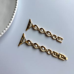 May include: Gold-toned watch band with a chain-link design. Each link features a small, round, white pearl. The band is designed to attach to a smartwatch. The background is a plain white surface.