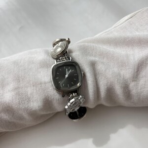 May include: Silver wristwatch with a square, dark-coloured face and a linked band. The band features decorative circular and floral elements. The watch is displayed on a white surface.