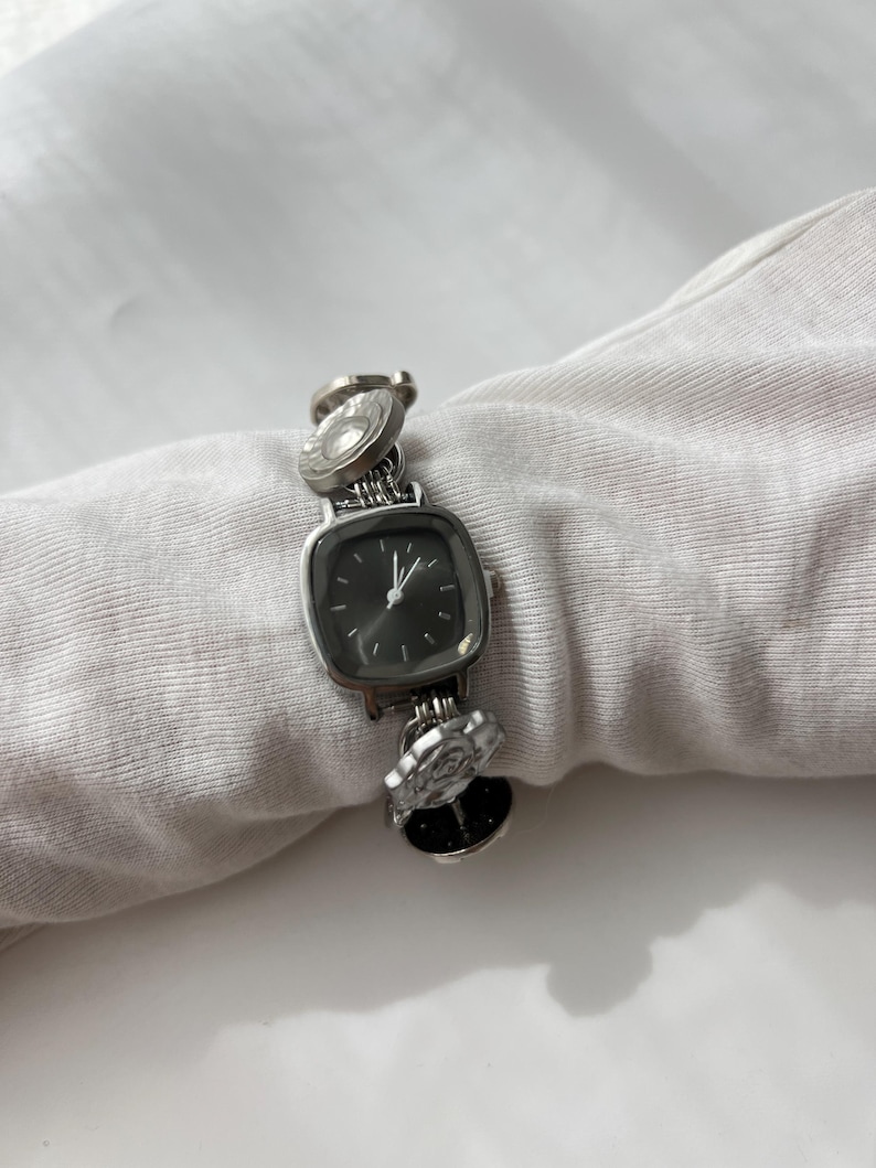 May include: Silver wristwatch with a square, dark-coloured face and a linked band. The band features decorative circular and floral elements. The watch is displayed on a white surface.