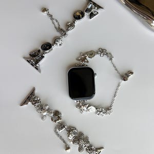 May include: A silver-toned smartwatch with a black screen and two decorative watch bands. One band features black circular charms and butterfly accents, while the other displays silver floral motifs, small pearls, and dangling heart-shaped charms.