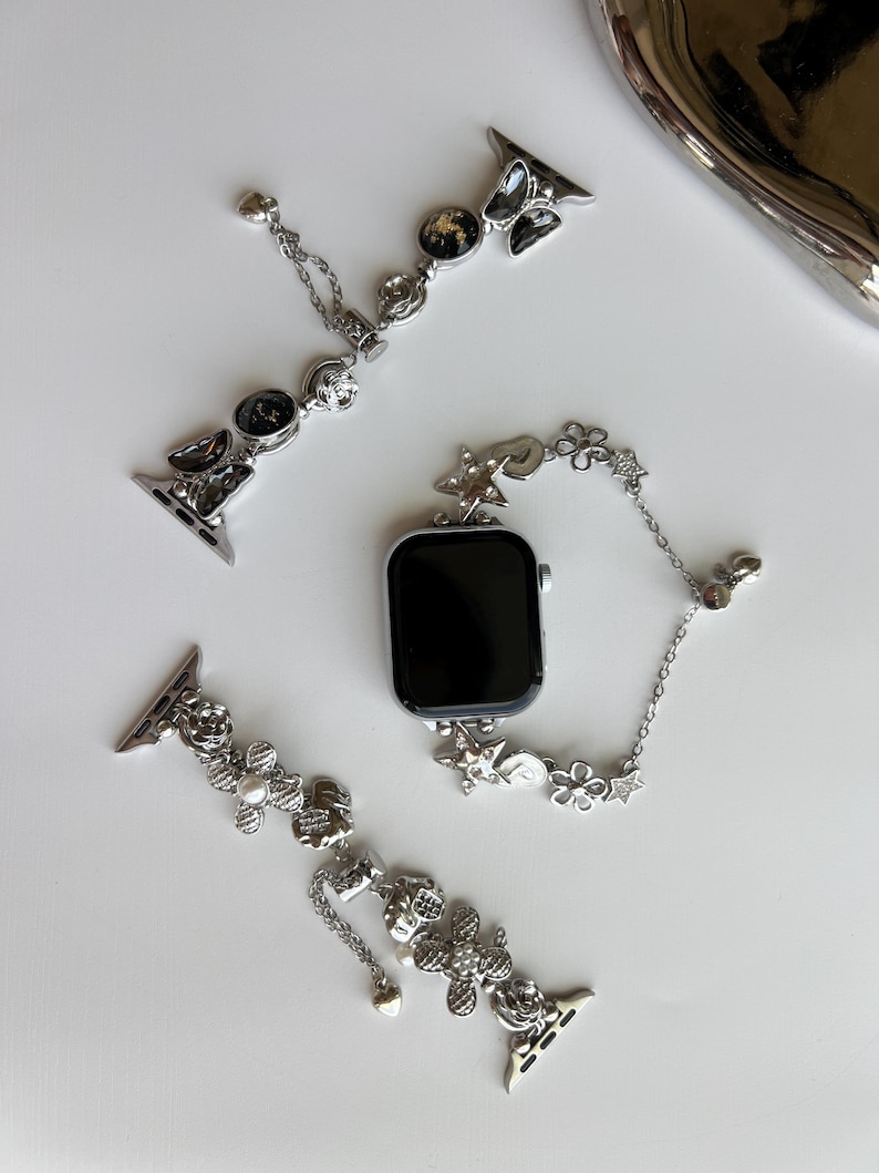 May include: A silver-toned smartwatch with a black screen and two decorative watch bands. One band features black circular charms and butterfly accents, while the other displays silver floral motifs, small pearls, and dangling heart-shaped charms.