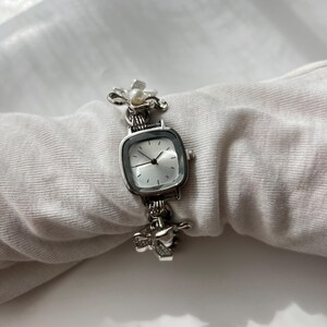 May include: Silver wristwatch with a square face and a light-coloured dial. The watch features decorative floral and heart-shaped accents on the band. A small pearl is set in the floral design. The watch is displayed on a white fabric.