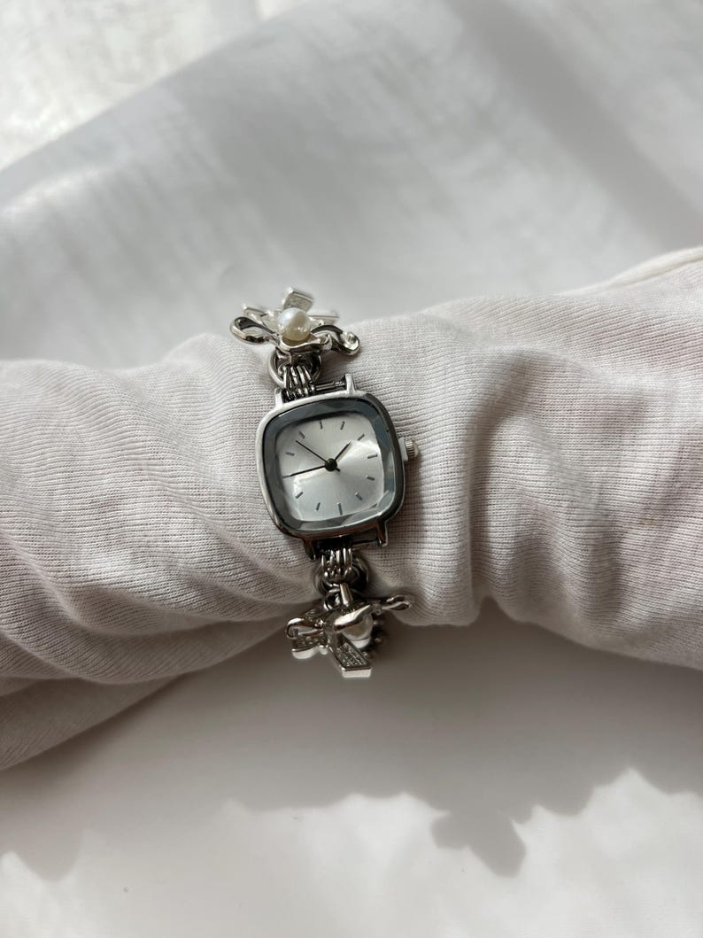 May include: Silver wristwatch with a square face and a light-coloured dial. The watch features decorative floral and heart-shaped accents on the band. A small pearl is set in the floral design. The watch is displayed on a white fabric.