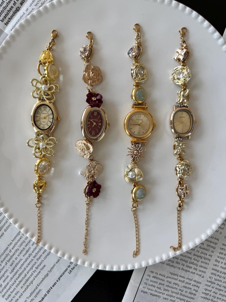 May include: Four gold-toned bracelet watches with decorative charms and oval or round faces are displayed on a white plate. The watches have different colored faces and embellishments, including flowers and pearls.