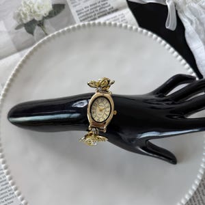 May include: A gold-toned wristwatch with a cream-colored face and black numerals. The watch features a floral design on the band and around the face. The watch is displayed on a black hand-shaped display.