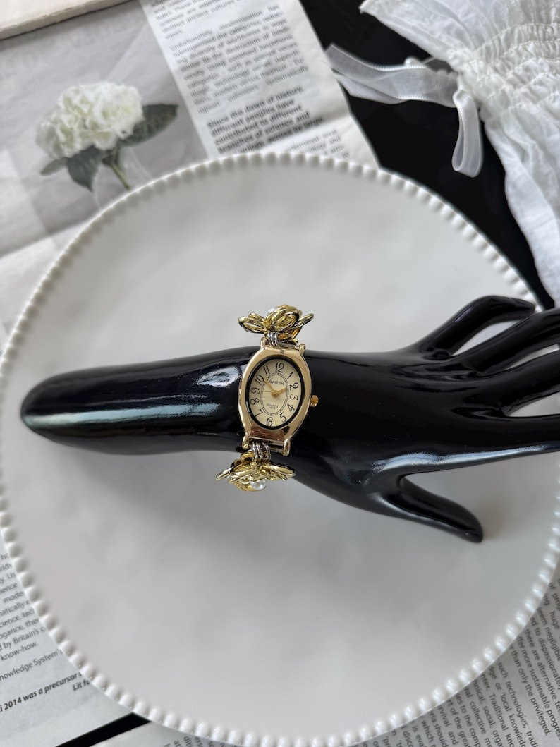 May include: A gold-toned wristwatch with a cream-colored face and black numerals. The watch features a floral design on the band and around the face. The watch is displayed on a black hand-shaped display.