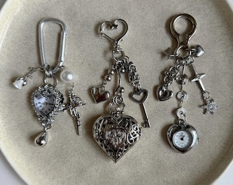 May include: Three silver-toned keychains with heart-shaped pendants and decorative charms. One keychain features a small watch, another a locket, and the third a key. The keychains are displayed on a cream-colored plate.