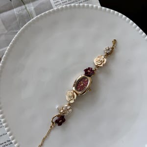 May include: Gold-toned watch with a burgundy oval face and floral accents. The watch band features gold flowers, burgundy floral clusters, and a small pearl. The watch is displayed on a white plate.