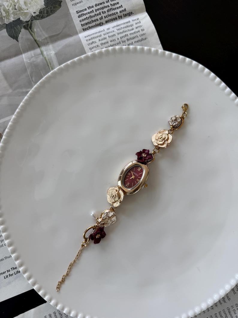 May include: Gold-toned watch with a burgundy oval face and floral accents. The watch band features gold flowers, burgundy floral clusters, and a small pearl. The watch is displayed on a white plate.