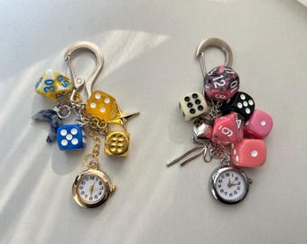 May include: Two keychains with dice and pocket watches. One keychain has yellow, blue, and gold dice, a gold watch, and a silver clasp. The other has pink, black, and white dice, a silver watch, and a silver clasp.