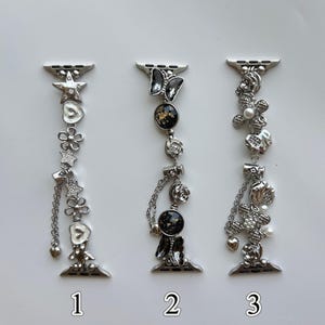 May include: Three silver-toned watch bands featuring decorative charms. The first band has stars, flowers, and hearts. The second band features a butterfly, circular black and gold accents, and roses. The third band displays floral motifs with pearls.