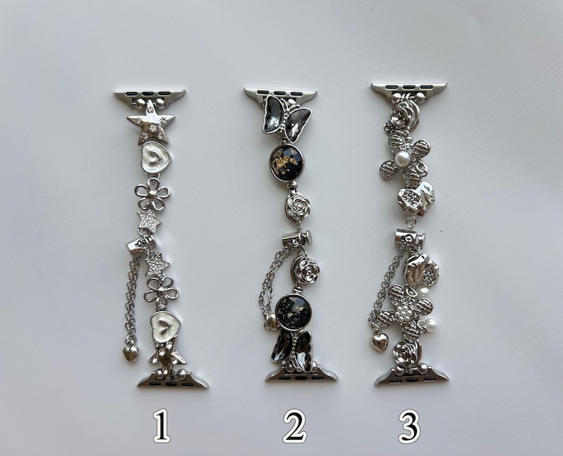 May include: Three silver-toned watch bands featuring decorative charms. The first band has stars, flowers, and hearts. The second band features a butterfly, circular black and gold accents, and roses. The third band displays floral motifs with pearls.