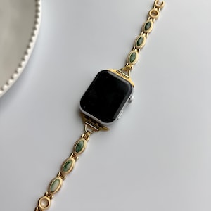 May include: Gold-toned watch band with oval green stone accents. The band is connected to a silver smartwatch with a black screen. The watch band features a clasp closure and a decorative design.