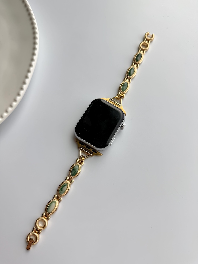 May include: Gold-toned watch band with oval green stone accents. The band is connected to a silver smartwatch with a black screen. The watch band features a clasp closure and a decorative design.