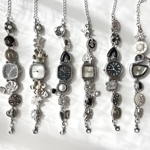 Może przedstawiać: Sześć srebrnych zegarków z zawieszkami z kwadratowymi i okrągłymi tarczami. Każdy zegarek posiada ozdobne zawieszki, w tym kwiaty, serca i gwiazdy. Zegarki wiszą na srebrnych łańcuszkach na białym tle.