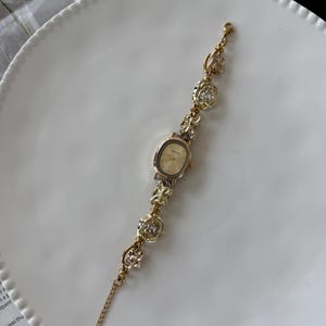 May include: Gold-toned wristwatch with an oval face and a delicate chain bracelet. The bracelet features decorative floral and geometric charms with clear crystal accents. The watch is displayed on a white plate.