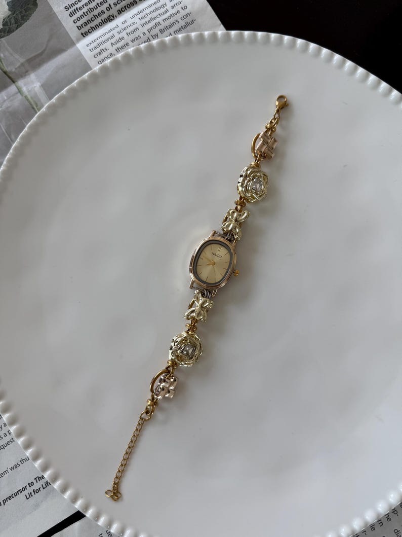 May include: Gold-toned wristwatch with an oval face and a delicate chain bracelet. The bracelet features decorative floral and geometric charms with clear crystal accents. The watch is displayed on a white plate.