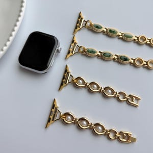 May include: A silver smartwatch with a black screen is next to three gold-colored watch bands. Two bands have green oval stones, and two have white pearls. The bands are designed to attach to the watch.