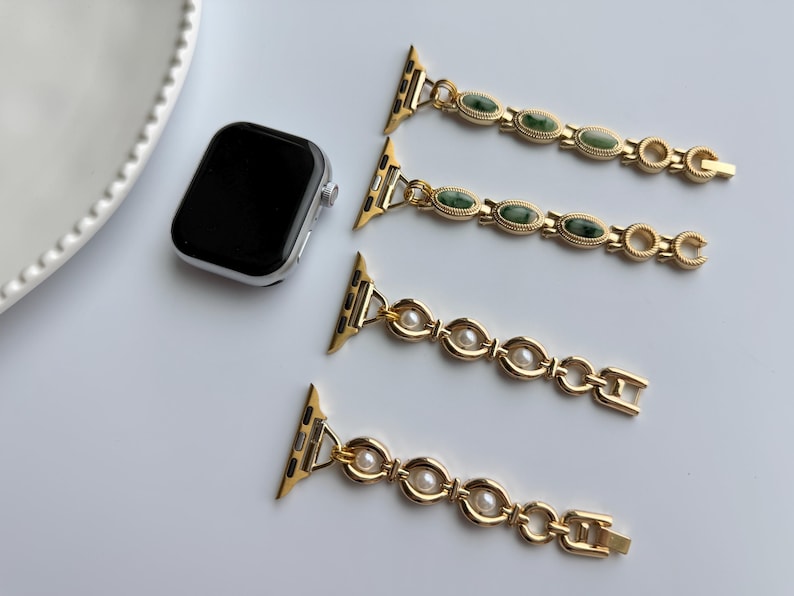 May include: A silver smartwatch with a black screen is next to three gold-colored watch bands. Two bands have green oval stones, and two have white pearls. The bands are designed to attach to the watch.