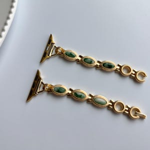 May include: Two gold-toned watch bands with green oval-shaped stones. The bands have a decorative design with a clasp closure. The watch bands are displayed on a white surface.