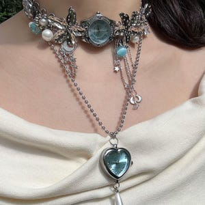 Peut inclure: Un collier de montre argenté avec un cadran bleu clair. Le collier présente un style ras du cou avec des accents floraux et perles, des chaînes pendantes et un pendentif en forme de cœur avec une goutte de perle.