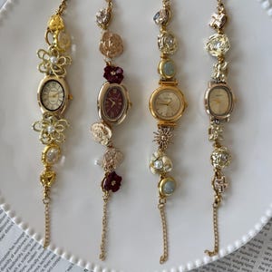 May include: Four gold-toned bracelet watches with decorative charms and oval or round faces are displayed on a white plate. Each watch has a different design, including floral and gem accents, and a delicate chain.