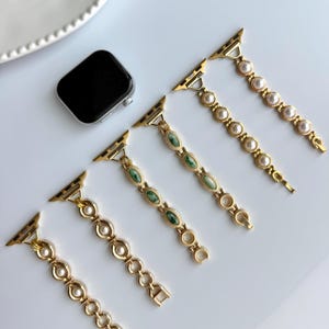 May include: Gold-toned watch bands with pearl and green stone accents. The bands are displayed with a black smartwatch on a white surface. The bands feature a variety of designs, including oval green stones and round pearls.